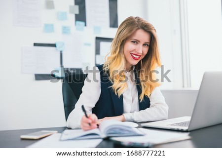 young blond smiling businesswoman works in her laptop in modern office, taking notes to her business planner,looks confident and busy, multitasking, work concept Royalty-Free Stock Photo #1688777221