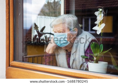lonely senior woman with surgical mask sitting on a window plane at home, coronavirus and covid-19 provisions Royalty-Free Stock Photo #1688780245