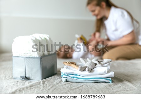 Mom caresses her baby, in the foreground diapers, clothes and toys suitable for age