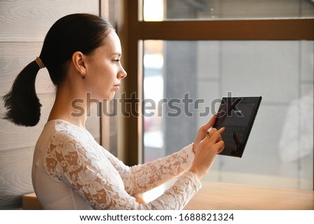 girl works in a cafe with a tablet, enters a password, hairstyle tail, against the background of a window