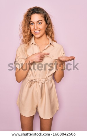 Young beautiful woman with blue eyes wearing casual summer dress over pink background amazed and smiling to the camera while presenting with hand and pointing with finger.