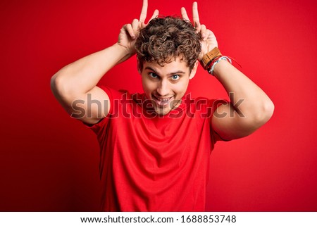 Young blond handsome man with curly hair wearing casual t-shirt over red background Posing funny and crazy with fingers on head as bunny ears, smiling cheerful