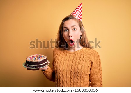 Young beautiful blonde woman holding birthday cake over isolated yellow background scared in shock with a surprise face, afraid and excited with fear expression