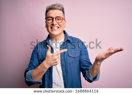 Young handsome modern man wearing glasses and denim jacket over pink isolated background amazed and smiling to the camera while presenting with hand and pointing with finger.