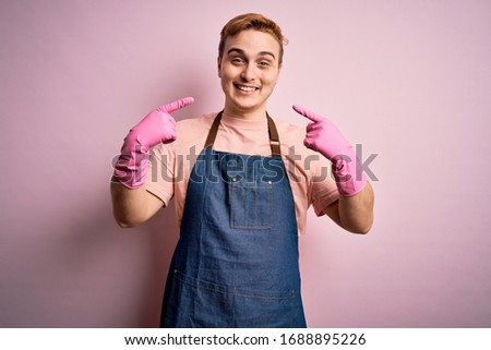 Young handsome redhead cleaner man doing housework wearing apron and gloves smiling cheerful showing and pointing with fingers teeth and mouth. Dental health concept.