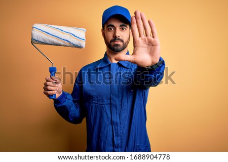 Young handsome painter man with beard wearing blue uniform and cap painting using roller with open hand doing stop sign with serious and confident expression, defense gesture