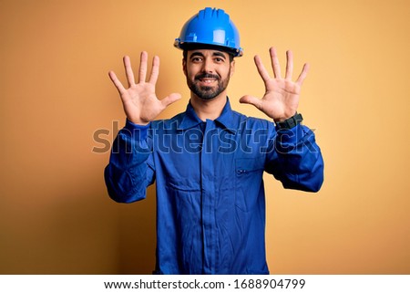 Mechanic man with beard wearing blue uniform and safety helmet over yellow background showing and pointing up with fingers number ten while smiling confident and happy.