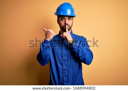 Mechanic man with beard wearing blue uniform and safety helmet over yellow background asking to be quiet with finger on lips pointing with hand to the side. Silence and secret concept.