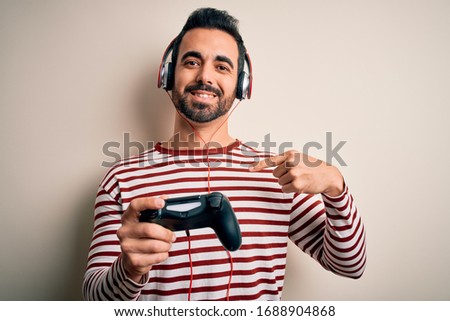 Young handsome gamer man with beard playing video game using joystick and headphones with surprise face pointing finger to himself