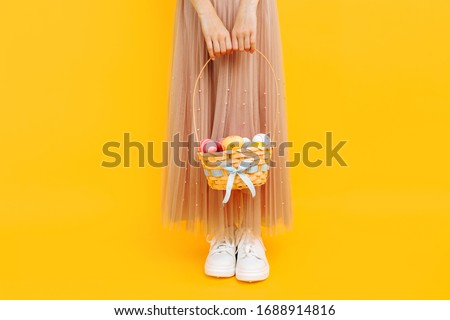 Close-up photo of legs, a girl holding a basket with Easter eggs on a yellow background. Symbol of Easter and spring