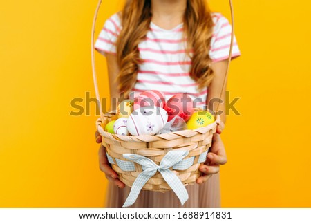 A beautiful happy girl on a head with rabbit ears, holding a wicker basket with colorful eggs on a yellow background. Symbol of Easter and spring