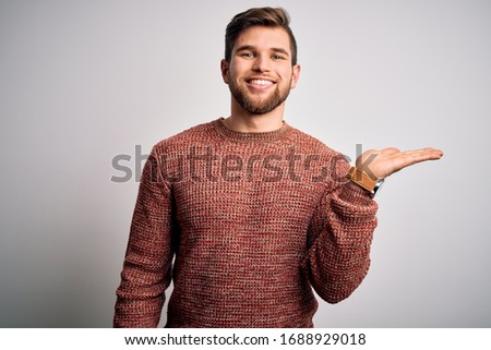 Young blond man with beard and blue eyes wearing casual sweater over white background smiling cheerful presenting and pointing with palm of hand looking at the camera.
