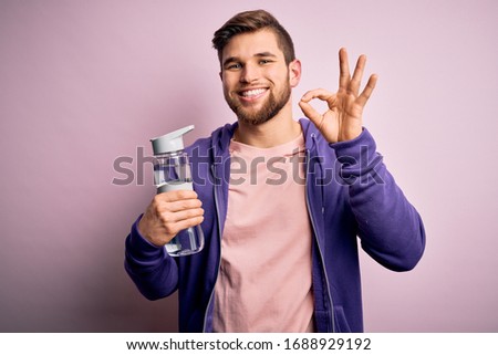 Young blond sporty man with beard and blue eyes doing sport holding bottle of water doing ok sign with fingers, excellent symbol