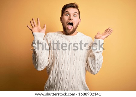 Young blond man with beard and blue eyes wearing white sweater over yellow background celebrating crazy and amazed for success with arms raised and open eyes screaming excited. Winner concept