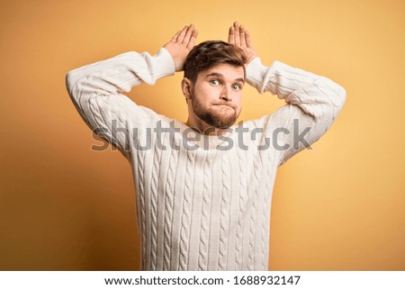 Young blond man with beard and blue eyes wearing white sweater over yellow background Doing bunny ears gesture with hands palms looking cynical and skeptical. Easter rabbit concept.
