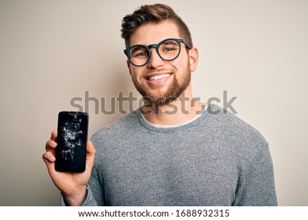 Young blond man with beard and blue eyes wearing glasses holding broken smartphone with a happy face standing and smiling with a confident smile showing teeth
