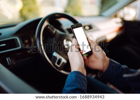 Hand holds the cell phone against the background of the steering wheel of the car. Phone with a blank  screen for an inscription or picture. Transportation and vehicle concept. 