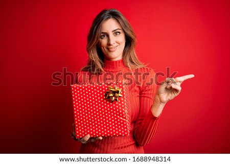 Young beautiful brunette woman holding birthday gift over isolated red background very happy pointing with hand and finger to the side