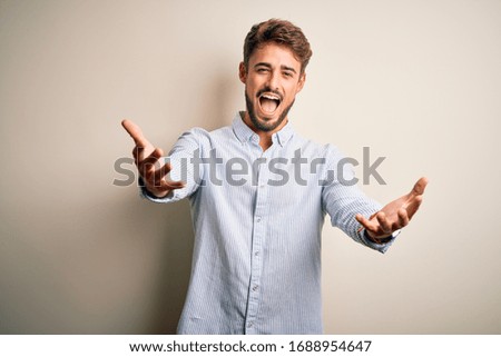 Young handsome man with beard wearing striped shirt standing over white background smiling cheerful offering hands giving assistance and acceptance.