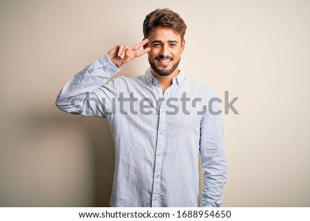 Young handsome man with beard wearing striped shirt standing over white background Doing peace symbol with fingers over face, smiling cheerful showing victory