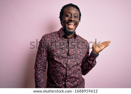 Young handsome african american man wearing casual shirt standing over pink background smiling cheerful presenting and pointing with palm of hand looking at the camera.