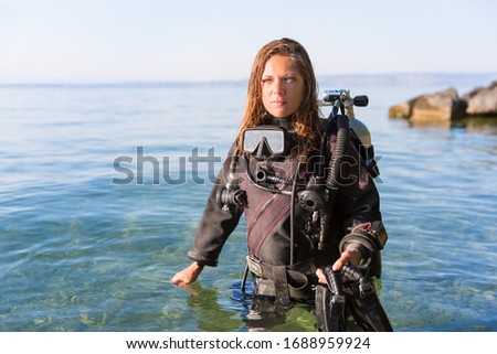 Female Scuba Diving Instructor Standing in Water Wearing a Dry Suit, a Twin Tank and Holding Fins Royalty-Free Stock Photo #1688959924