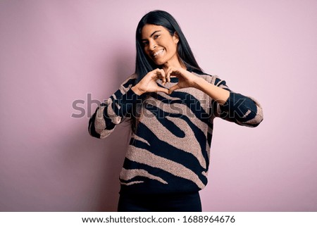 Young beautiful hispanic woman wearing animal print sweater over pink background smiling in love showing heart symbol and shape with hands. Romantic concept.