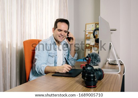 Boy photographer answering the phone while working from home on his computer. On the table are an electronic graphics tablet and a photo camera