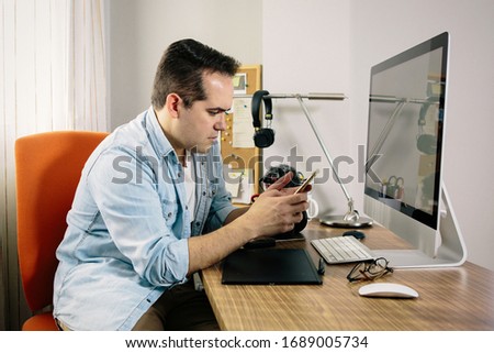 Boy photographer attending to messages on his phone while working from home on his computer. On the table are an electronic graphics tablet and a photo camera.