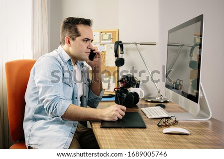 Boy photographer answering the phone while working from home on his computer. On the table are an electronic graphics tablet and a photo camera
