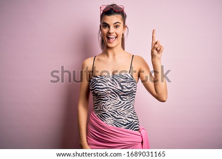 Young beautiful brunette woman on vacation wearing swimsuit over pink background pointing finger up with successful idea. Exited and happy. Number one.