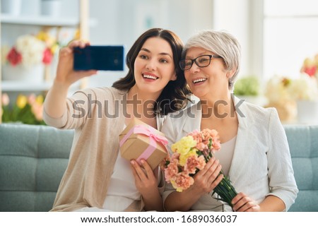 Happy mother's day! Beautiful young woman and her mother with flowers and gift box at home. 