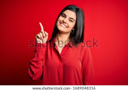 Young beautiful brunette woman wearing casual shirt standing over red background showing and pointing up with finger number one while smiling confident and happy.