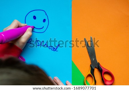 Home school and a smiley face written by a child on blue paper. With a childs hand holding a pen and scissors. Left handed.