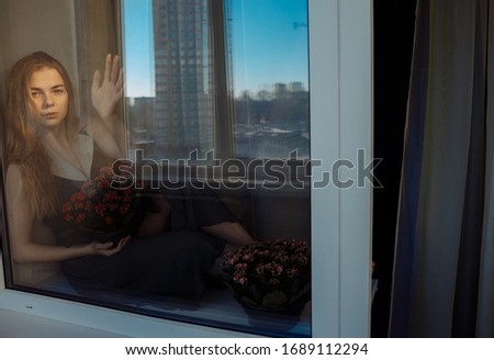 Home isolation. The girl is sitting at home by the window. Outside the window, blue sky and houses. In the arms of the girl are flowers. The girls hand is pressed into the glass.