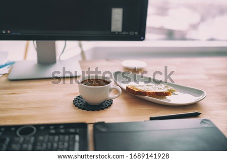 Working from home office remote work breakfast snack coffee and toast on table next to keyboard, pc screen, drawing digital tablet for freelance retouching, graphic design worker.