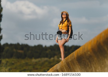 Delighted slim female travel photographer enjoying her work standing outdoors with backpack camera at neck in casual denim shorts yellow t-shirt hat sunglasses.