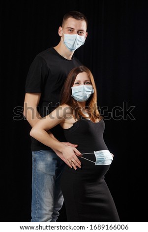 pregnant woman in a black dress with dark hair and a man  with a medical mask against coronavirus pandemic infection COVID-19 on a black background. Little girl holds a mask near the abdomen