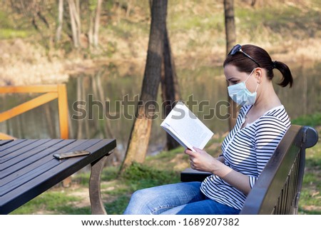 Woman with a protective mask on her face, reading a book sitting on a bench in city park by the lake.