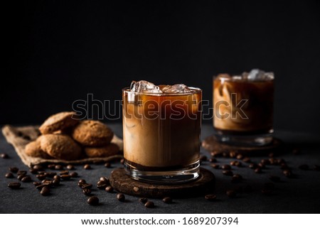 A horizontal photo of 2 rocks glasses with cold iced coffee with milk, coffee beans around, cookies, dark background, deep shadows