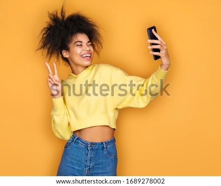 Young girl holds smartphone and takes selfie photo, smiles at camera. Photo of african american girl on yellow background