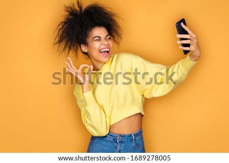Young girl takes selfie photo and makes okay gesture, smiles at camera. Photo of african american girl on yellow background
