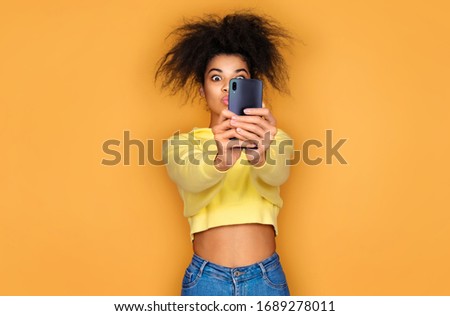 Young girl holds smartphone and takes selfie photo. Photo of african american girl on yellow background