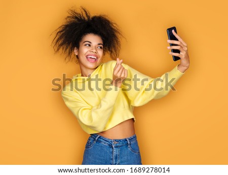 Young girl makes korean love sign and takes selfie photo, looking at phone. Photo of african american girl on yellow background