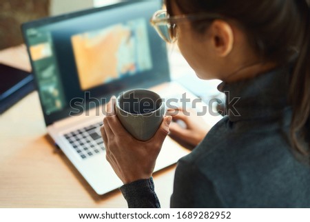 Rear view woman freelancer typing on laptop while sitting at deskr. Skilled graphic designer drinking coffee and working on a new project. Remote work, freelance, using laptop computer. focus on cup
