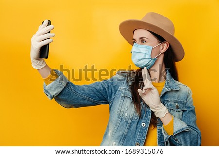 Brunette woman holding phones in hands makes selfie, against yellow background, dressed in denim jacket, wears protective medical mask to protect herself from coronavirus. Social Distancing concept