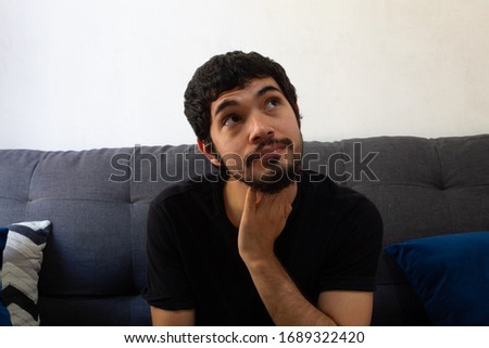 Bearded Hispanic young man sitting on a sofa very thoughtful looking out a window