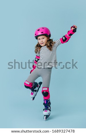 Serious focused little girl in striped clothes and roller skates with pink protective gear over blue background. She's showing off for a photo, standing on one leg, holding other one with her hand.