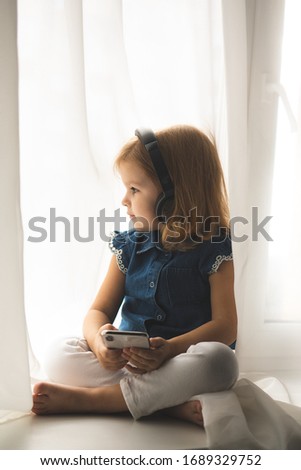 Beautiful little caucasian girl in a casual listening to the music in a headphones and watching cartoons on smartphone or phone. Baby girl use headphones and hold the phone. 