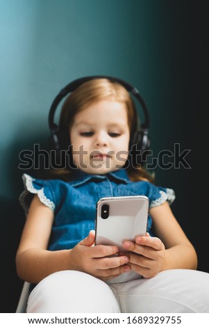 Beautiful little caucasian girl in a casual listening to the music in a headphones and watching cartoons on smartphone or phone. Baby girl use headphones and hold the phone. 